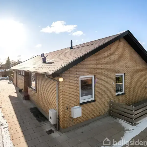 En gul murstensbygning står på en flisebelagt sti; lidt sne dækker jorden. Bygningen har vinduer og et lavt plankeværk i forgrunden. Himlen er solrig med få skyer.