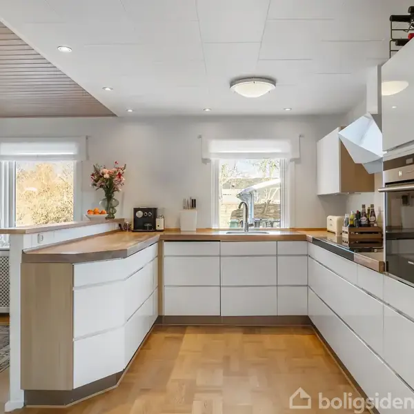 Køkken har hvide moderne skabe, en indbygget ovn og komfur, ovenlys og parketgulv. Blomster og frugtskål pryder bordet ved siden af en lys spiseplads med vinduer bagved.