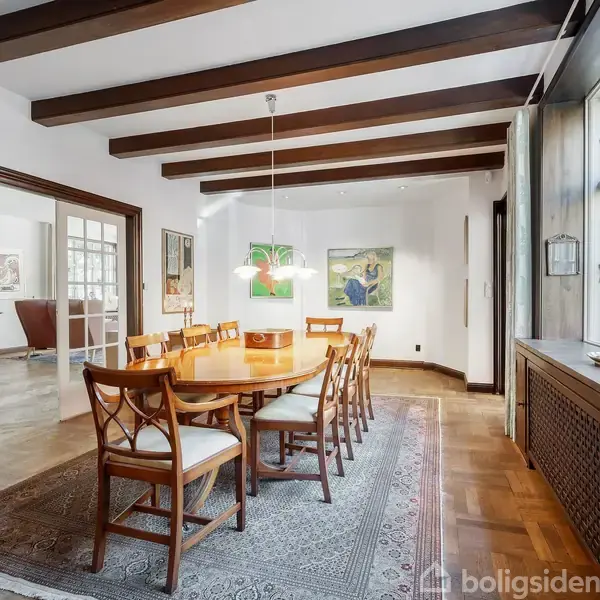 Spisebord med otte stole står på et tæppe i spisestuen. Træbjælker i loftet, malerier på væggen. Store vinduer til højre giver naturligt lys.
