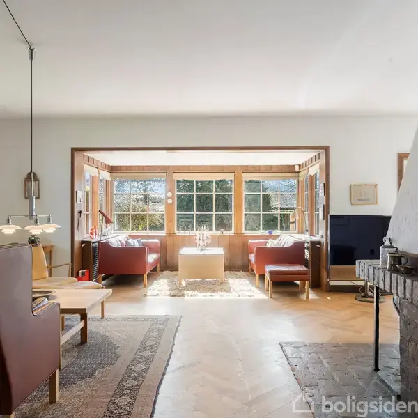 Stue med polstrede sofaer og bord, badet i sollys, ved store vinduer. Stuen har et trægulv og en pejs til højre, indrettet med billeder på væggen og lamper.