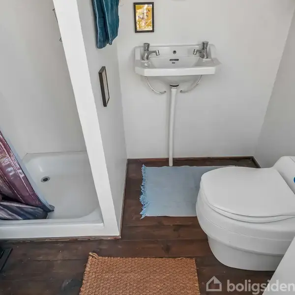 Toilet er placeret ved siden af en lille håndvask på et badeværelse med hvidt vægdesign. Brusekabinen med farverigt forhæng ligger ved siden af. Gulvet har et brunt trægulv.