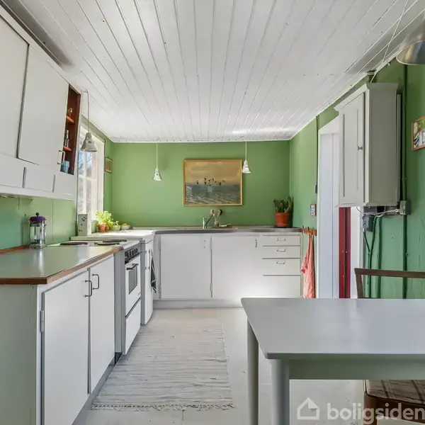 Køkken med hvide skabe og træbordplade, hvor der står en kedel. En bordlampe hænger over bordet. Grønne vægge med et maleri og vinduer ved siden af et bord.