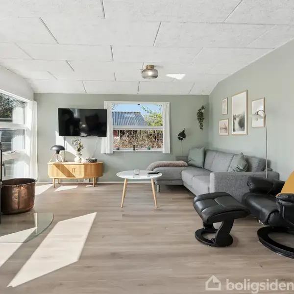 Stue med grå sofa og sort lænestol i moderne indretning. Brændeovn i hjørnet ved hoveddør, fladskærms-TV på væggen, og vindue med udsigt til træer. Stuebord og dekorationer.