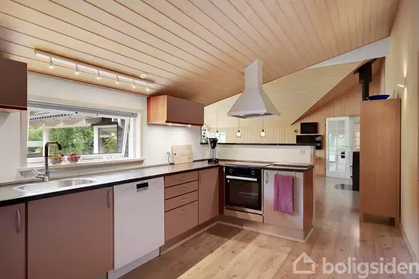 Køkken med lyse træelementer, kogeplade og emhætte, opvaskemaskine og store vinduer til venstre. Rummet er lyst med en skrående loft, og træplanker dækker gulv og loft.