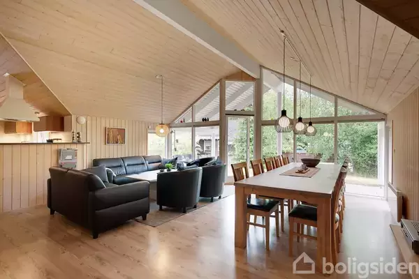Stue med sorte sofaer står ved store vinduer. Et langt spisebord med stole er oplyst af hængelamper. Trævægge og lofter skaber en hyggelig atmosfære i lyse omgivelser.