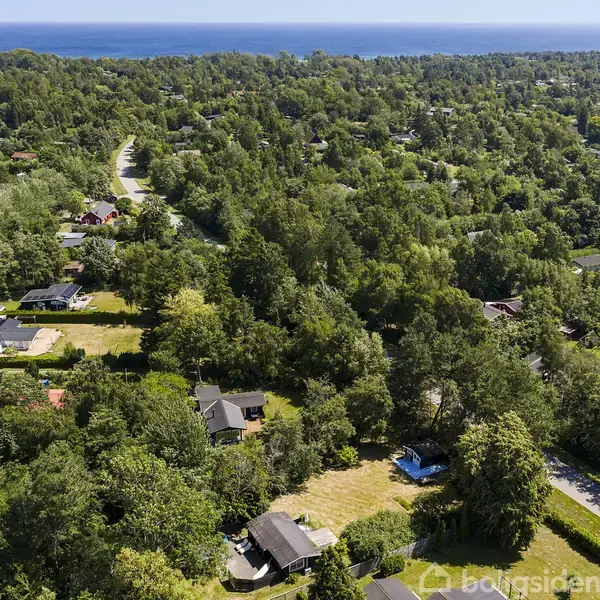 Huse ligger spredt blandt frodig skov og grønne træer. En vej snor sig gennem landskabet, og i horisonten ses havet. Boligområdet er fredfyldt og naturskønt.