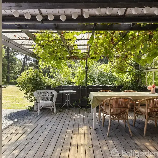 Terrassemøbler står under en pergola med grønne klatreplanter. Trebordet er dækket med dug, omgivet af flettede stole. Omkring er der frodig have med græsplæne og forskellige buske.