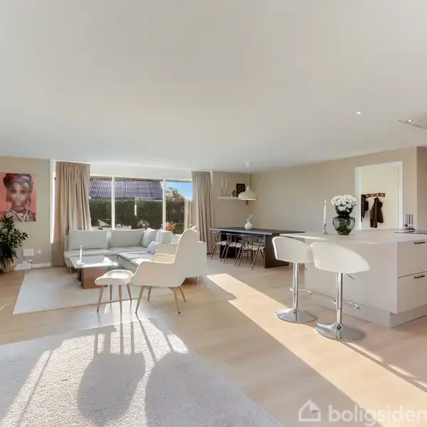 En lys, moderne stue med sofa og lænestole ved vindue. En åben køkkenø med barstole står til højre. Sollys strømmer ind gennem store vinduer. Møblerne er minimalistiske.