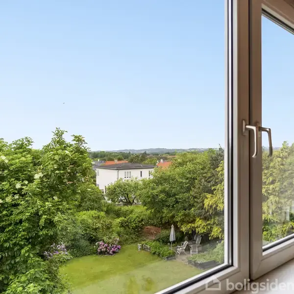 Et vindue åbent mod en frodig have med buske, træer og blomster. På vindueskarmen står flasker og dekoration. Udsigten viser også et rødt hustag i det fjerne.