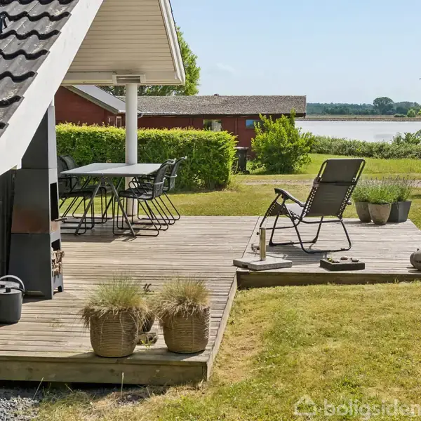 Træterrasse med en solvogn og cafébord i en have nær en sø. Solvognen står afslappet. Omkring ses en græsplæne, buske og udsigt til vandet med grønne omgivelser.