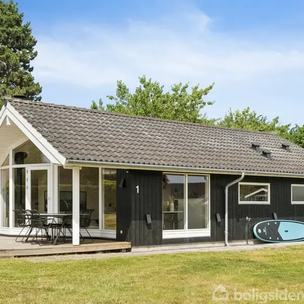 Sommerhus med sort træfacade står stille i frodig have. Terrasse med havemøbler ligger foran et stort glasparti. Surfbræt læner sig op ad huset. Træer omgiver området.
