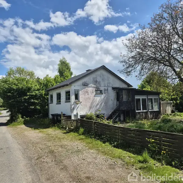 Et ældre, hvidt hus med sort tag står ved siden af en stille vej. Det har falmede vægge og er omgivet af træer og buske under en blå himmel med skyer.