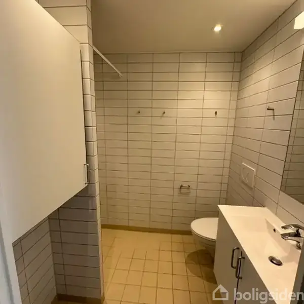 Et enkelt badeværelse har hvide fliser på væggene og gulfarvede fliser på gulvet. En vask og et toilet er placeret langs højre væg, med kroge på den bagerste væg.
