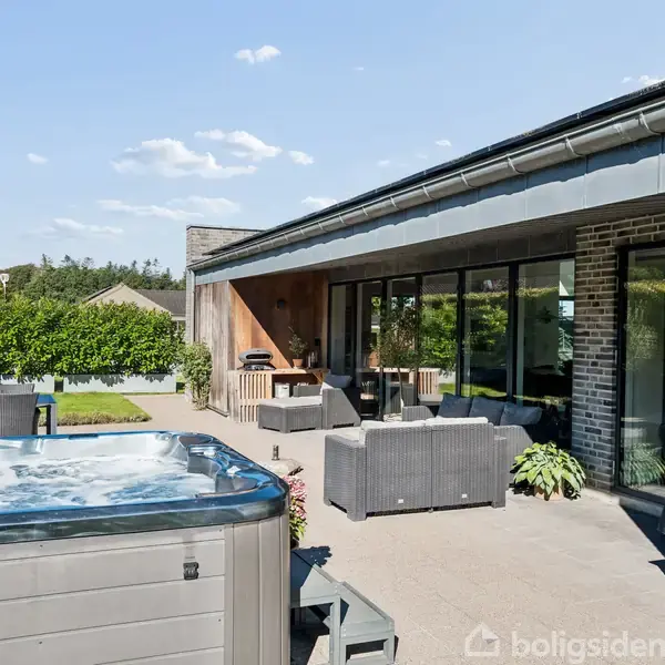 Udendørs terrasse med spabad i forgrunden. Loungeområde med sofa og grill. Omkringliggende have med grønne planter under en blå himmel. Moderne hus i baggrunden med store vinduer.