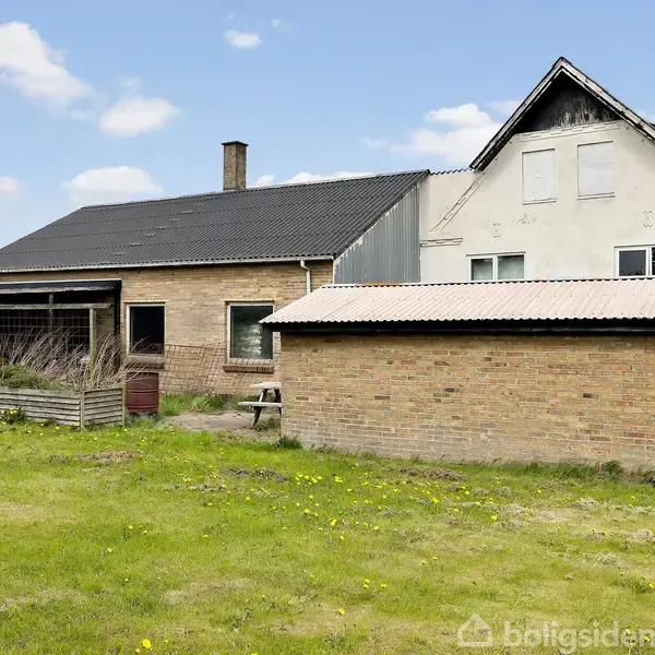 Hus i mursten med sort tag og mindre tilbygning. Står på grøn græsplæne med blå himmel og få skyer.