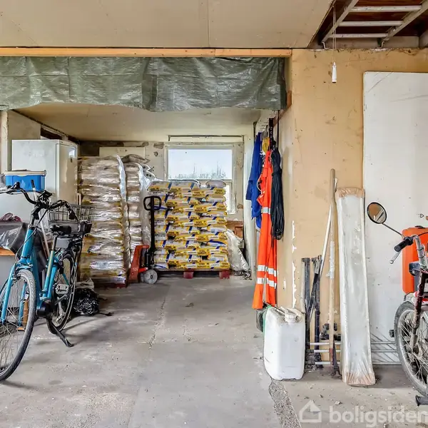 Cykel placeret i garage, omgivet af værktøj, lagersække og arbejdsudstyr. En motorcykel står til højre ved siden af opbevaringshylder. Hylderne indeholder hjelme og diverse materialer.