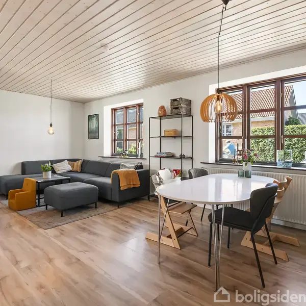 Stue med hjørnesofa i gråt, arrangeret rundt om et lille bord, ved siden af et spisebord med stole. Trægulve og lofter, store vinduer med udsigt til have.