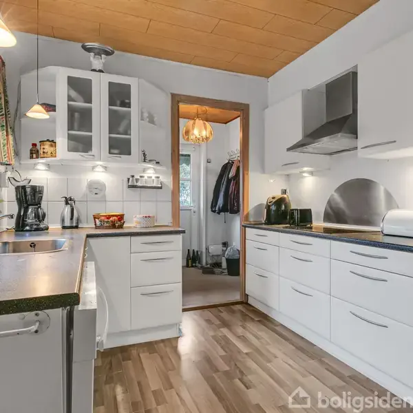 Et hvidt køkken med træbordplader, kaffemaskine og elkedel på bordet. Skabe er åbne med porcelæn indeni. Store vinduer og lamper hænger fra loftet.