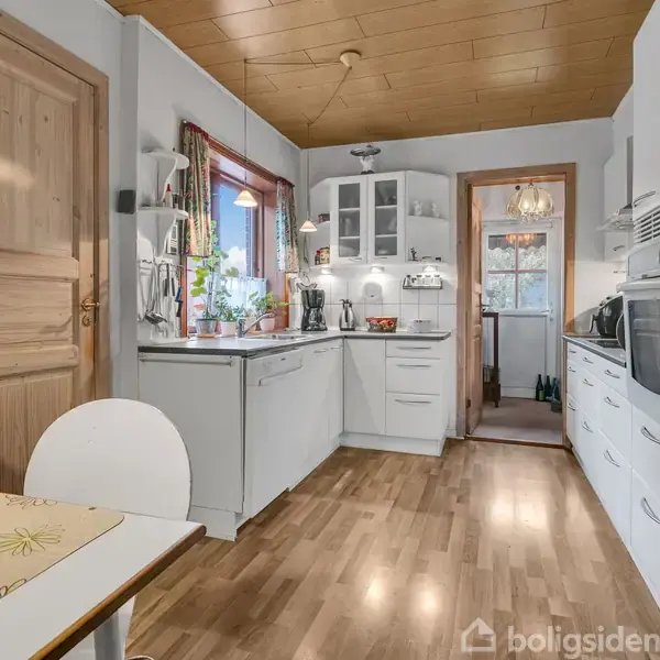 Et køkken viser hvide skabe og apparater, herunder en ovn og mikrobølgeovn, med et spisebord i forgrunden. Trægulv og -loft skaber en varm atmosfære.