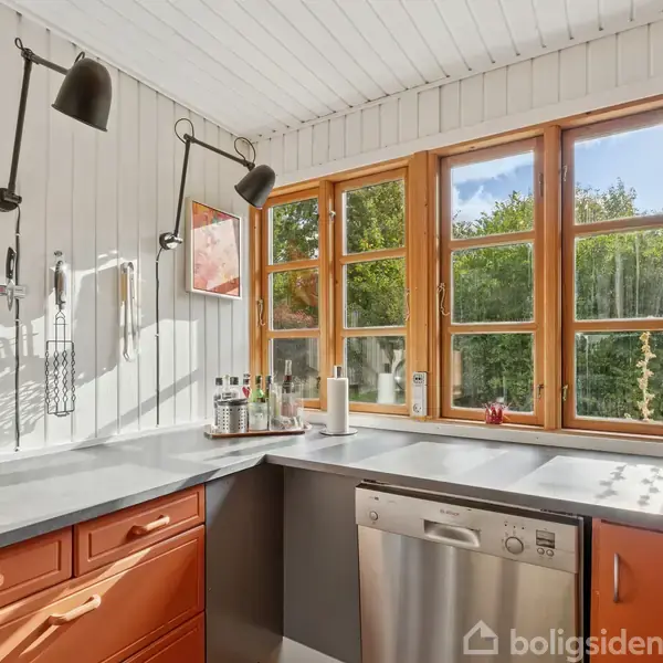 Et køkken med orange skabe og en opvaskemaskine under en grå bordplade. På væggen hænger køkkenredskaber. Store vinduer slipper lys ind og viser en grøn udsigt.