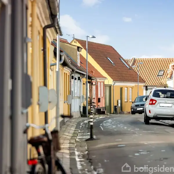 En hvid bil kører ned ad en smal, brostensbelagt gade flankeret af farverige, ældre bygninger med røde tegltage. Det er solskin, og en cykel står op ad en bygning.