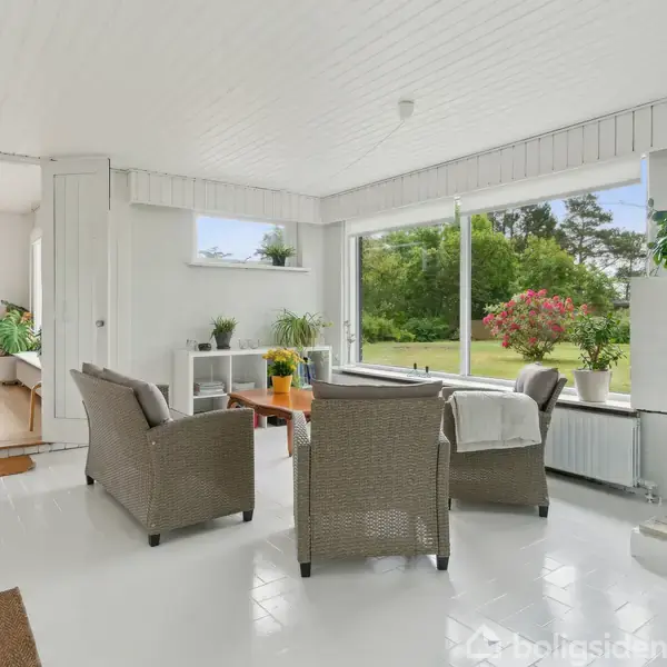 Stue med rottingmøbler, brændende pejs og stort vindue, der viser en grøn have med blomster. Lyse, hvide vægge og gulve skaber et lyst rum fyldt med planter.