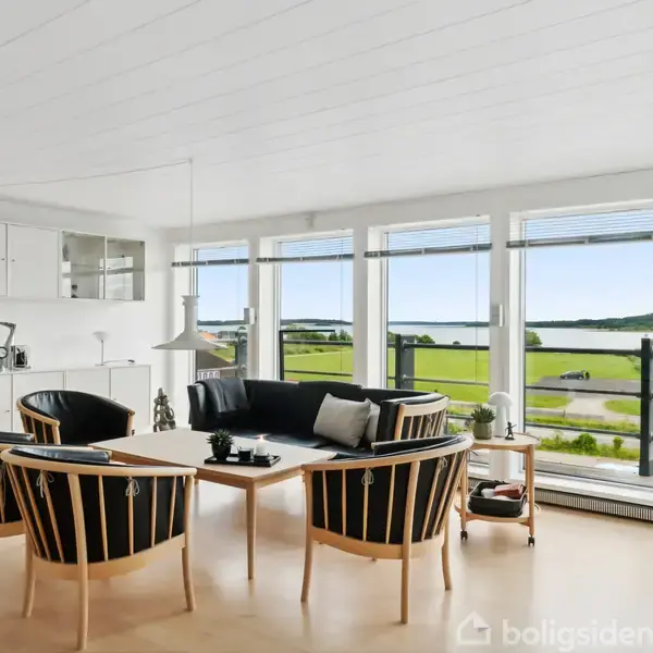 En pejs i sort murværk står på en hvidmalet væg. To moderne lænestole er placeret i et lyst rum med store vinduer, der giver udsigt til en terrasse og planter.
