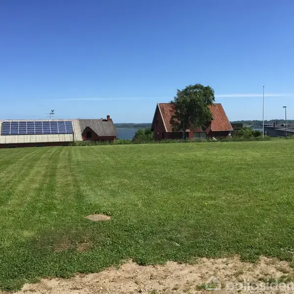 En stor græsplæne strækker sig i forgrunden; i baggrunden står to huse, det ene med solpaneler på taget, omgivet af en klar, blå himmel og en sø i horisonten.