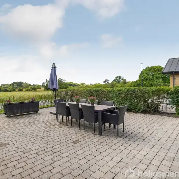 Terrasse med et bord og otte stole står på grå fliser. En parasol er ved siden af bordet, omgivet af grønne buske og en græsplæne, med åben udsigt til marker.