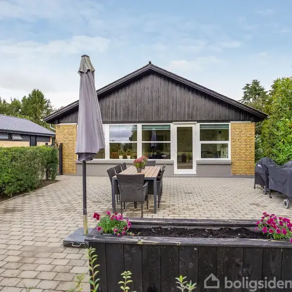 Hus i gule mursten med sort træbeklædt gavl. Terrasse med havebord, parasol og grill. Omkranset af grønne buske og træer. Blomsterkasse med pink blomster i forgrunden.