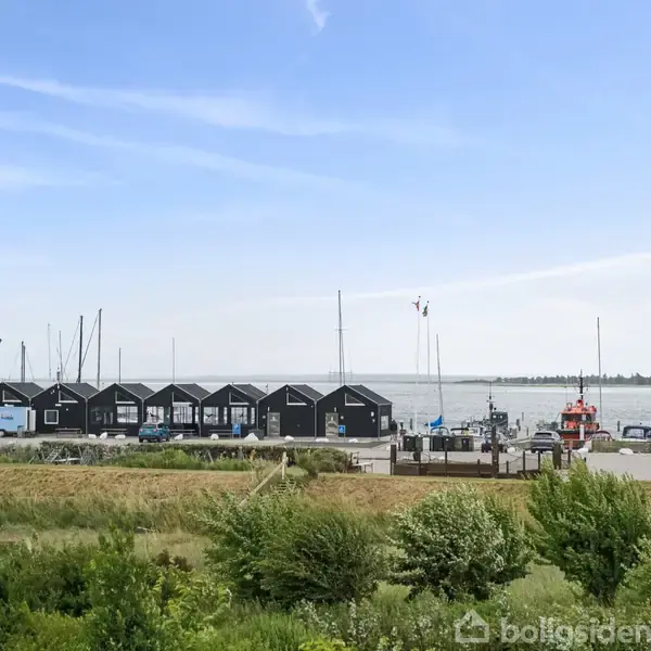 Både står stille ved kajen foran en række sorte skure ved en havn, omgivet af græs og buske, med havet i baggrunden under en klar blå himmel.