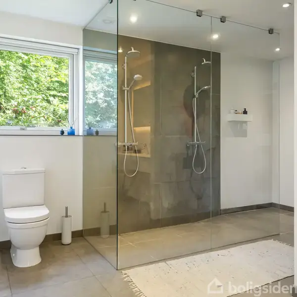 Toilet med hvide fliser står ved et vindue med grøn udsigt. Ved siden af er der en bruseniche med glasvæg og regnbruser i et lyst badeværelse.