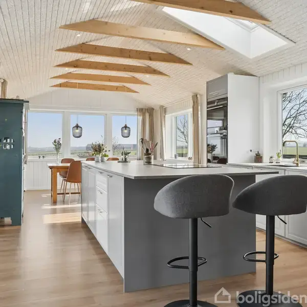 Et moderne køkken med grå køkkenø og to grå barstole. Sollys kommer ind fra loftsvinduer og store vinduer, hvilket skaber en lys atmosfære med udsigt til naturen udenfor.
