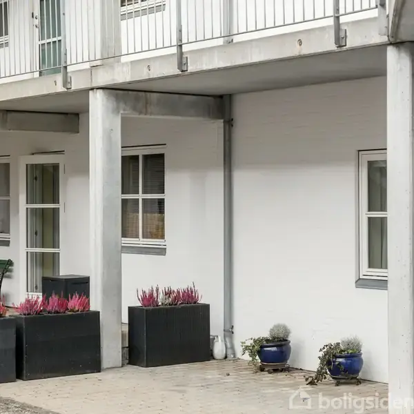 Balkon over skaber overdækning for terrassemøbler og plantekasser med lilla blomster ved hvid facade. Fliser dækker jorden. To vinduer og en dør ses på væggen.