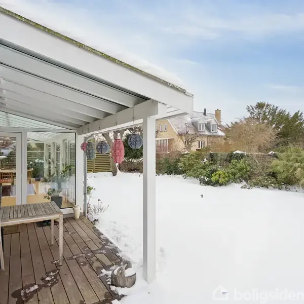 En overdækket terrasse med træbord og stole vender ud mod en snedækket have. Baggrunden viser huse og vinterlige træer under en klar himmel.