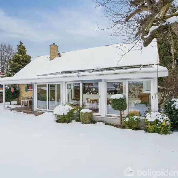 Et hus med store vinduer dækket af sne står i en vinterhave. Haven er dækket af et tykt lag sne, og der er træer og buske omkring huset.