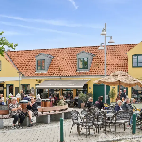 Folk sidder og står på en solrig plads foran et gult, klassisk hus med rødt tag og butikker. Rykind Murray står skrevet på facaden. Pladsen er omgivet af træer og parasoller.