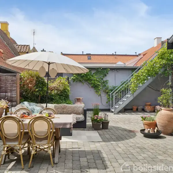 Terrasse med spisebord og stole, overdækket af en parasol. Sofa med puder står ved siden af. Omgivet af planter, trapper og mure i en solrig gårdhave.