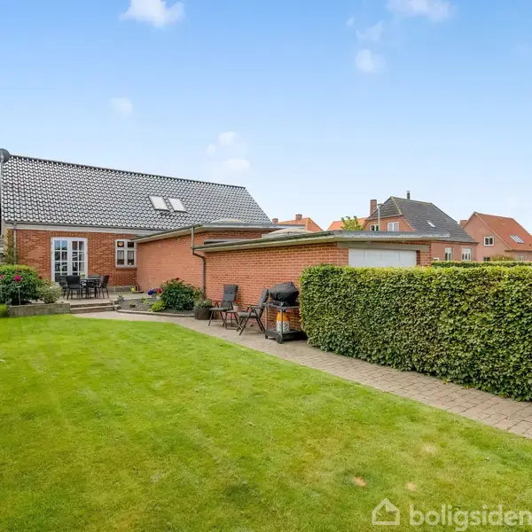 Rødt murstenshus med sort tag, omgivet af en grøn have med klippede hække og flisebelagt terrasse. Havemøbler står på terrassen. Andre huse ses i baggrunden under en blå himmel.