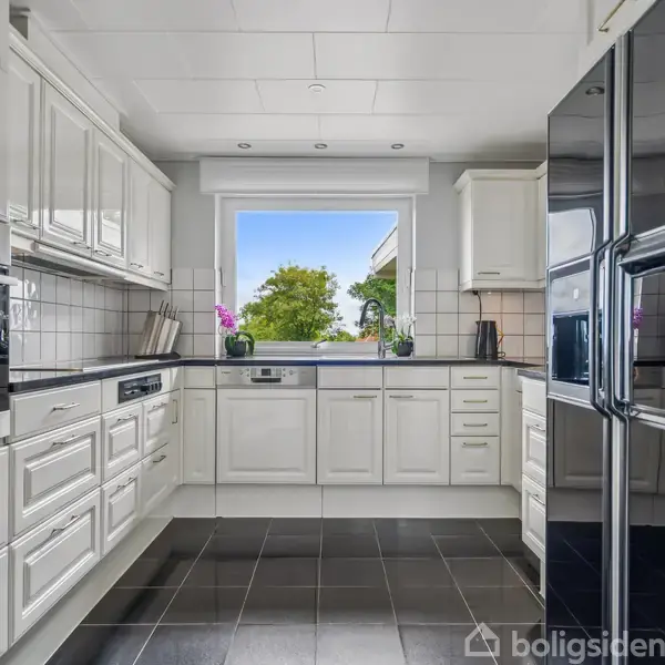 Et moderne køkken med hvide skabe og sorte bordplader. To ovne indbygget i væggen. Et stort vindue giver udsigt til en have. Indretningen er stilren og symmetrisk.