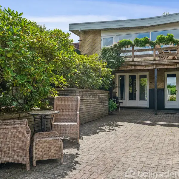 En terrasse med fliser har kurvestole og et bord. En overdækket balkon er fyldt med planter. Omgivet af murstensvægge og grønne buske, med en blå himmel ovenfor.
