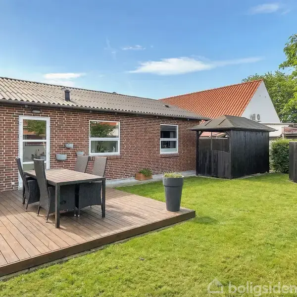 En træterrasse har et bord med fire stole, placeret ved en murstensbygning. En velholdt græsplæne omkranser området, og der står et sort skur til højre.