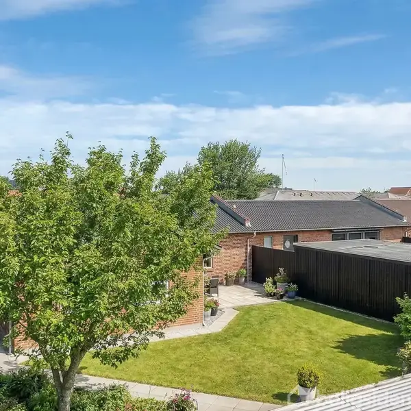Huset har røde mursten og sort tag, ligger i en velplejet have med grønne træer og en græsplæne, under en blå himmel. En indgang er synlig bag hækken.