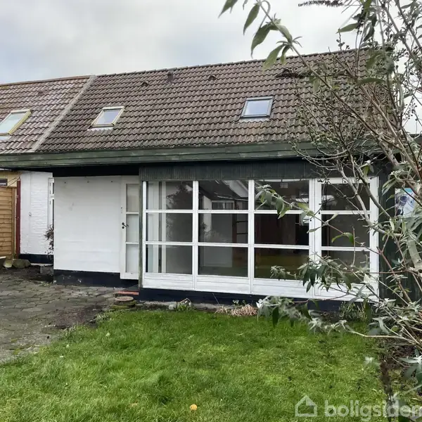 Et lille hvidt hus med en skråtag og ovenlysvinduer. Huset har en vinterhave med glaspartier. Det står i en have med græs og en stenbelagt terrasse.