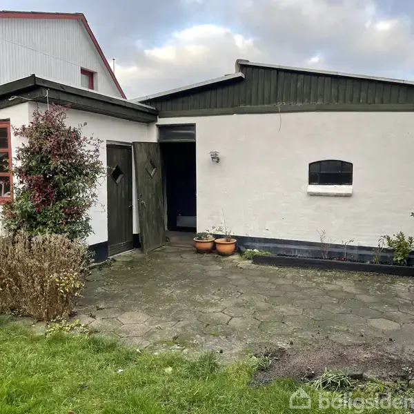 Et lille hvidt skur med sorte kanter står ved siden af et større hus. Døren er åben, og tre blomsterkrukker står foran. Det er omgivet af en flisebelagt gårdhave.