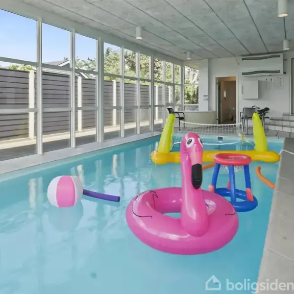 En indendørs pool er fyldt med forskellige oppustelige legetøj, herunder en pink flamingo og en strandbold. Omgivet af store vinduer med udsigt til en have udenfor.