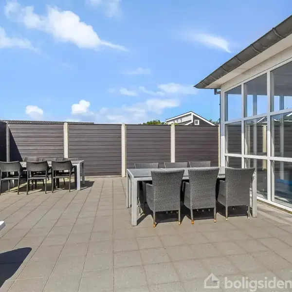 En udendørs terrasse med to spiseborde og flere stole. Terrassen har flisebelægning og er omgivet af højt hegn. Sollys stråler fra en klar blå himmel med få skyer.