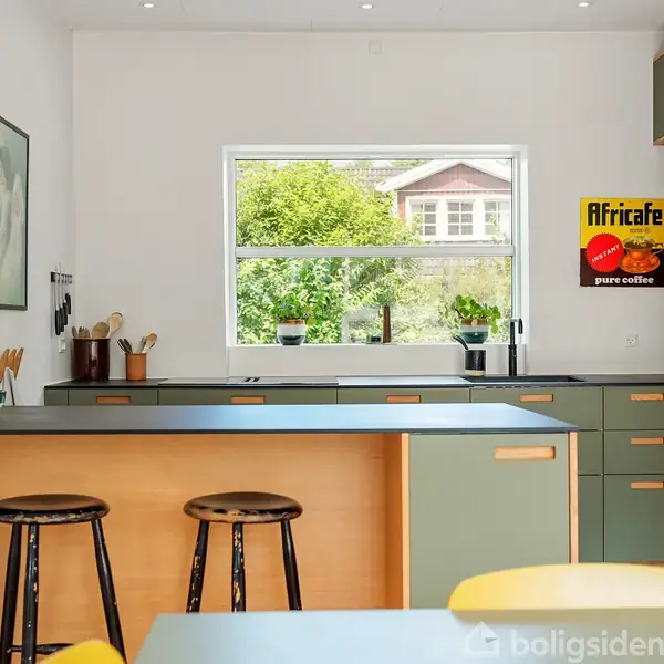 Køkken med to taburetter ved en ø, set mod et vindue med udsigt til have. Plakaten "Africaffe pure coffee" hænger på væggen, omgivet af grønne skabe og hylder.