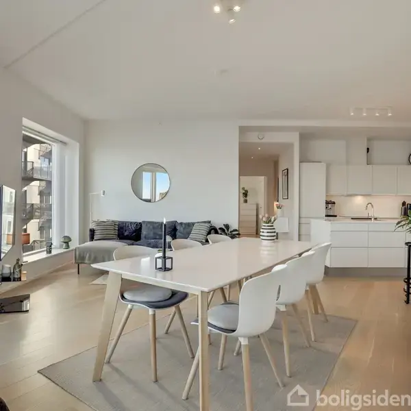 Spisebord med seks stole står på et lyst trægulv i en moderne stue. Stuen har hvide vægge, sofamøbler, et TV og et moderne køkken i baggrunden. Indretningen er minimalistisk.