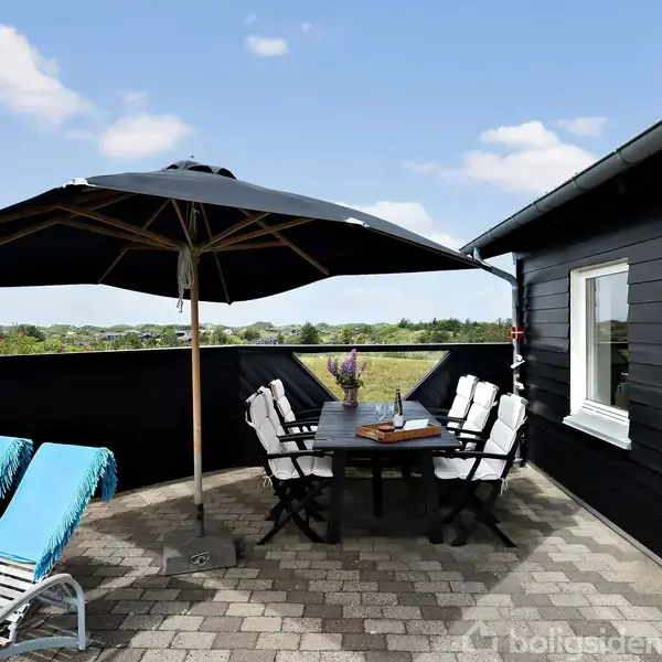 En terrasse med et sort havebord og seks stole placeret under en stor parasol. To liggestole med turkise hynder står ved siden af, omgivet af en hæk og blå himmel.